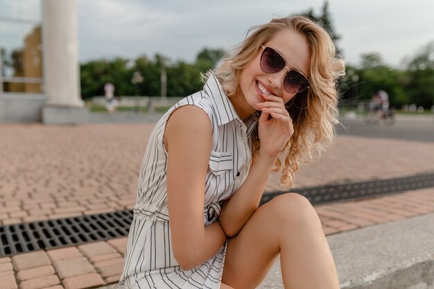 Young attractive cute stylish blonde woman sitting in city street in summer fashion style white cotton dress wearing sunglasses