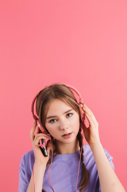 分離されたピンクの背景の上にカメラで思慮深く見ているヘッドフォンで音楽を聴いているライラックTシャツの2つの三つ編みを持つ若い魅力的な夢のような女の子