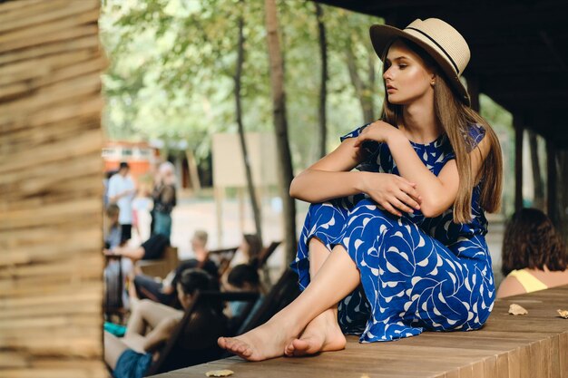 Молодая привлекательная мечтательная женщина в голубом платье и шляпе босиком задумчиво смотрит в сторону в городском парке