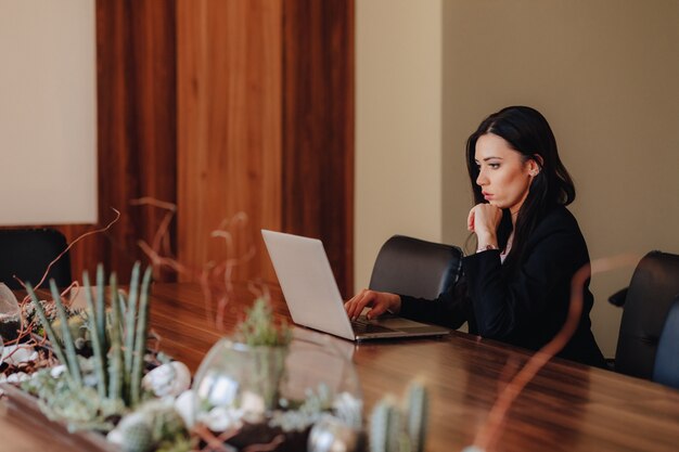 Young attractive emotional girl in business style clothes sitting at a desk on a laptop and phone in the office or auditorium