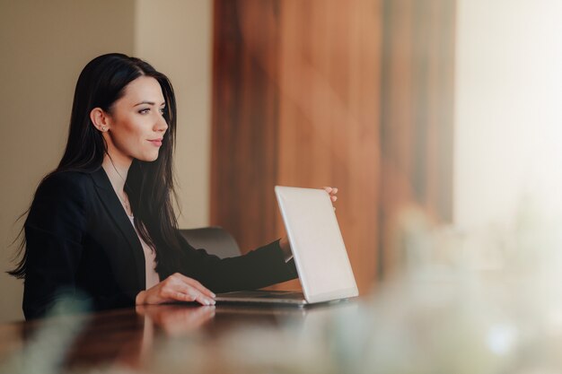 Young attractive emotional girl in business-style clothes sitting at a desk on a laptop and phone in the office or auditorium