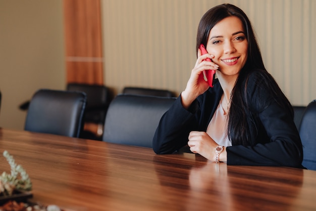 Free photo young attractive emotional girl in business style clothes sitting at desk with phone in office or audience