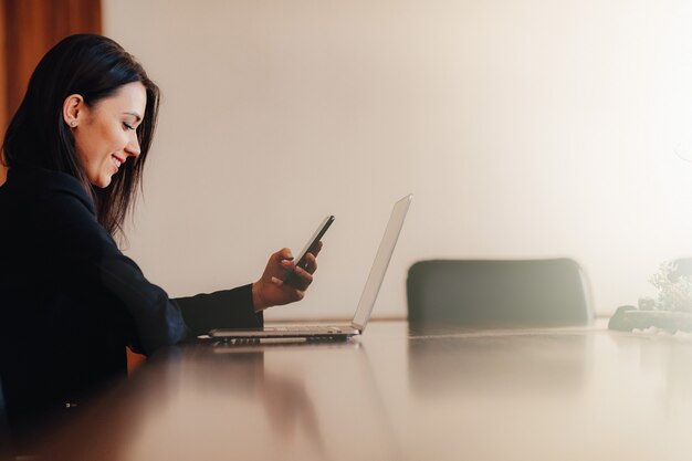 Young attractive emotional girl in business style clothes sitting at desk with phone in office or audience