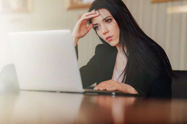 Young attractive emotional girl in businessstyle clothes sitting at a desk on a laptop and phone in the office or auditorium