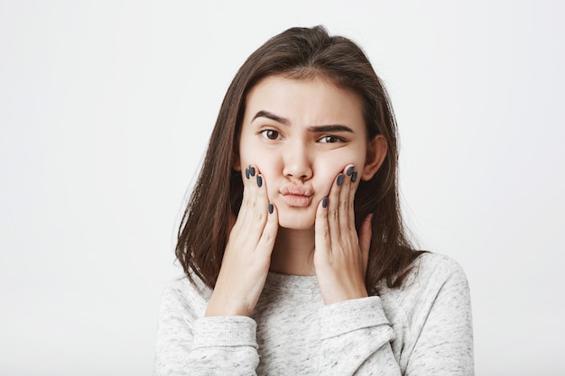 young attractive european female model squeezing her cheeks with puzzled and doubtful expression