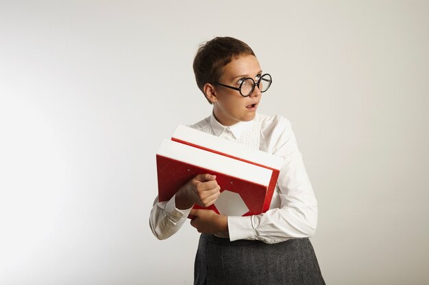 Young attractive European teacher in white blouse and gray skirt holding red and white binders rolls eyes in disbelief on white wall