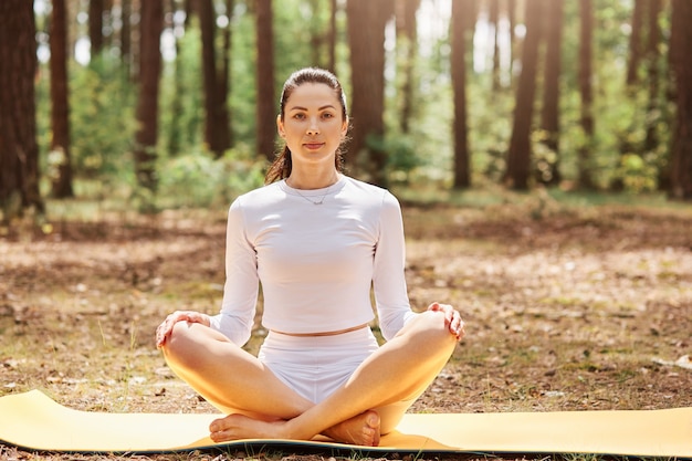 Young attractive female dresses stylish sportswear sitting with crossed legs on karemat,  keeps hands on knee, practicing yoga in forest.