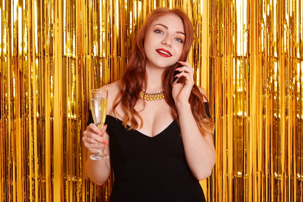 ワインやシャンパンのグラスを保持し、金色の見掛け倒しの壁に立っている黒のエレガントなドレスとネックレスを警告する若い魅力的な女性。