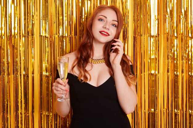 Молодая привлекательная женщина защищает черное элегантное платье и ожерелье, стоящее на золотой стене с мишурой, держа в руке бокал вина или шампанского.