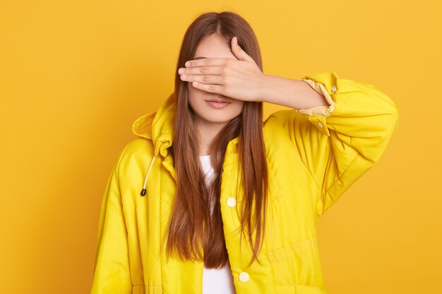 Young attractive female wearing jacket covering her eyes with hands, woman with long hair, standing against yellow wall, lady hides from her friend.