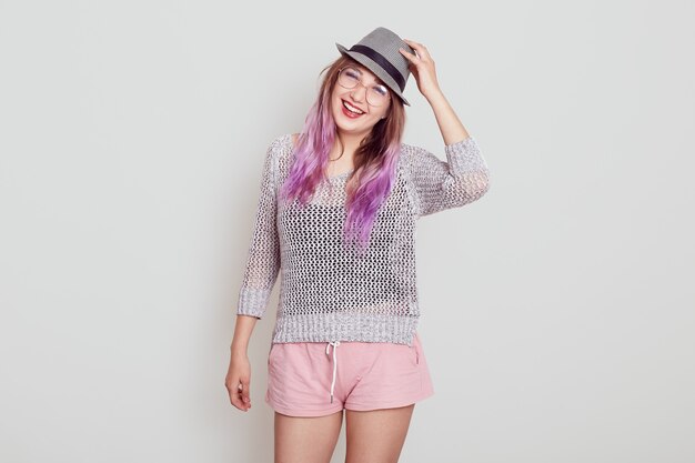 Young attractive female with happy facial expression looking directly at camera with toothy smile, touching her hat, being in good mood, isolated over grey background.