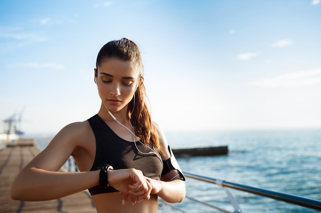 Foto gratuita giovane ragazza attraente fitness guardando l'orologio con la costa del mare sul muro