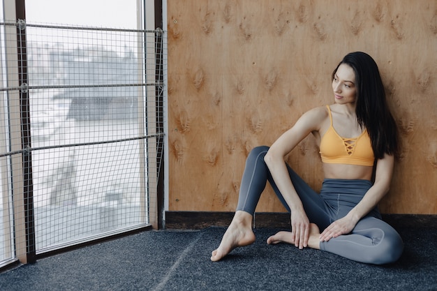 Young attractive fitness girl sitting on the floor near the window on the background of a wooden wall, resting on yoga classes