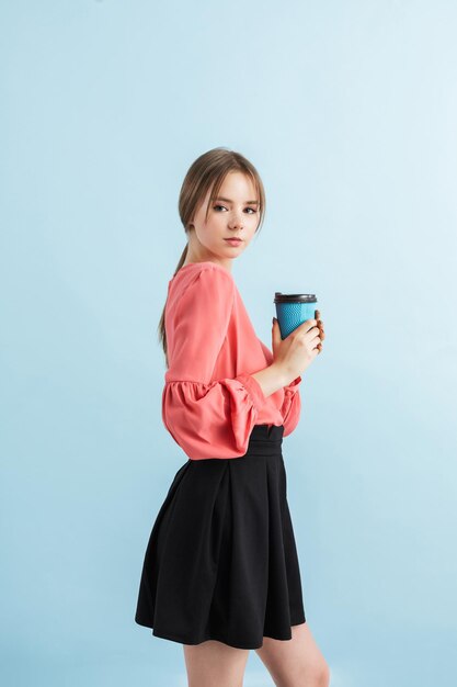 Молодая привлекательная девушка в блузке и черной юбке держит чашку кофе в руке, мечтательно глядя в камеру на синем фоне