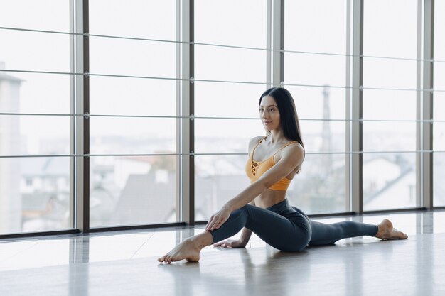 Young attractive girl doing fitness exercises with yoga on the floor against the surface of panoramic windows