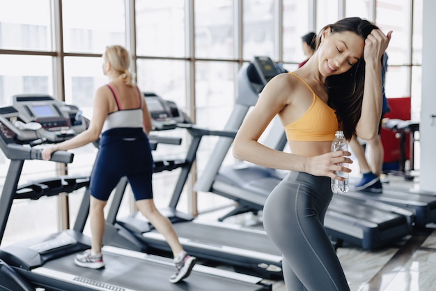 Young attractive girl in the gym drinking water on the background of treadmills