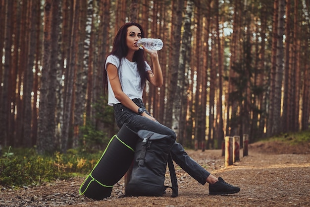 Молодая привлекательная девушка отдыхает в лесу и пьет воду.
