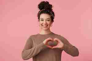Free photo young attractive girl with curly hair, sends a heart gesture, smiles broadly and looking at the camera isolated over pink background.