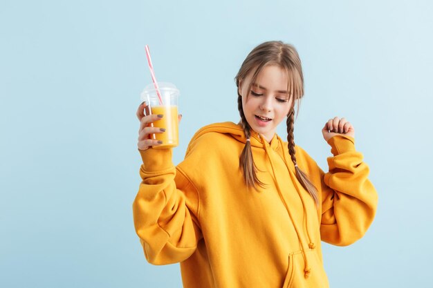 Giovane ragazza attraente con due trecce in felpa con cappuccio arancione che tiene in mano una tazza di plastica con succo di frutta mentre balla sognante su sfondo blu