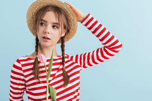 Young attractive girl with two braids in straw hat and red striped vest holding pink tulip in hand while dreamily looking aside over blue background