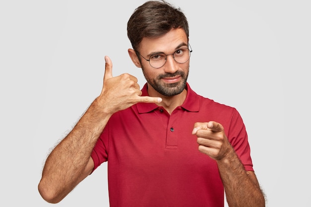 Young attractive guy in casual red t shirt, makes phone sign, indicates with index finger directly at camera