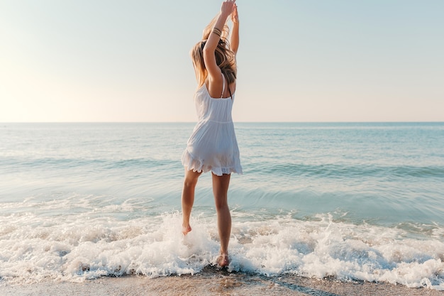 Молодая привлекательная счастливая женщина танцует поворачиваясь на берегу моря в солнечном летнем стиле моды в белом платье на каникулах