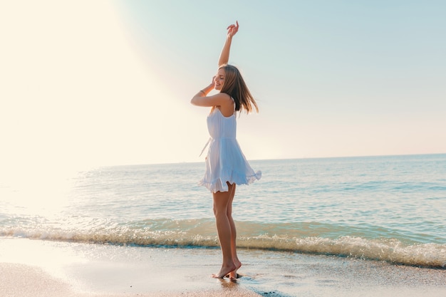 Молодая привлекательная счастливая женщина танцует, поворачиваясь на берегу моря в солнечном летнем стиле моды в белом платье