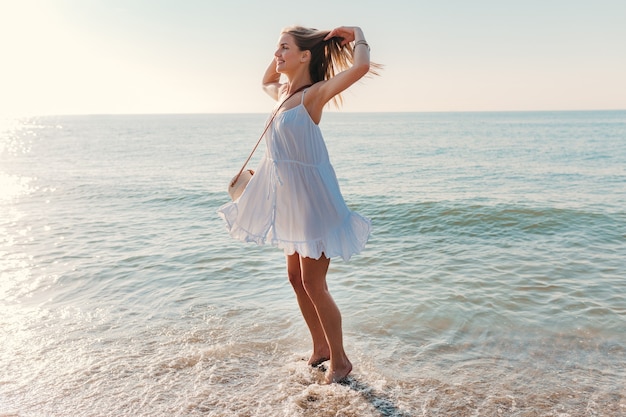 Молодая привлекательная счастливая женщина танцует, оборачиваясь на берегу моря в солнечном летнем стиле моды