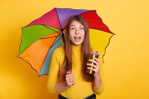 Young attractive lady with astonished facial expression posing with multicolored umbrella and coffee to go, stands with opened mouth