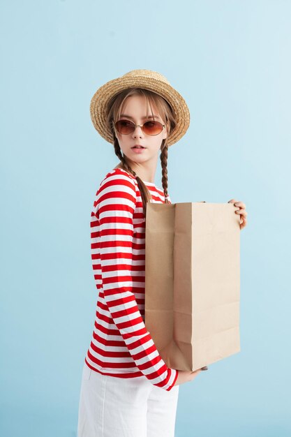麦わら帽子と赤いサングラスの2つのブレードを持つ若い魅力的な女性は、青い背景の上に紙袋を手に持っている間、思慮深く脇を見ています