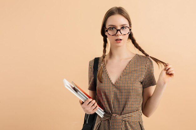 Young attractive lady with two braids in tweed jumpsuit and eyeglasses with black backpack on shoulder holding book and notepads in hand while dreamily looking in camera over beige background