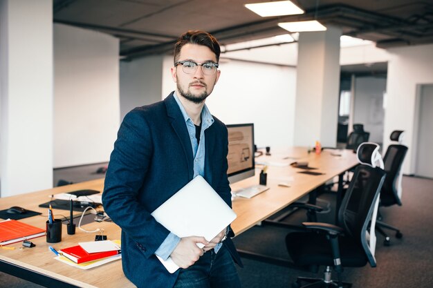 Young attractive man in glassess is standing near the workplace in office. He wears  blue shirt, dark jacket, laptop in hand. He is looking to the camera.