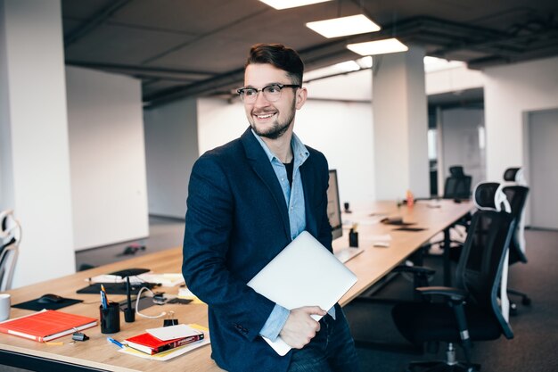 Young attractive man in glassess with beard  is standing near the workplace in office. He wears  blue shirt, dark jacket, laptop in hand. He is smiling to the side.