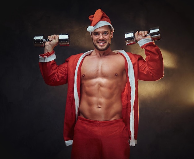 Free photo young attractive man in santa claus role is doing exercises with dumbbells.