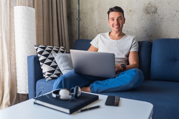 Young attractive man sitting on sofa at home working on laptop online, using internet