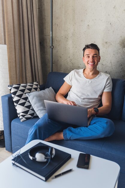 Young attractive man sitting on sofa at home working on laptop online