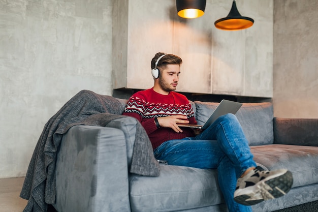 Young attractive man on sofa at home in winter in headphones, listening to music, wearing red knitted sweater, working on laptop, freelancer, serious, busy, typing, concentrated