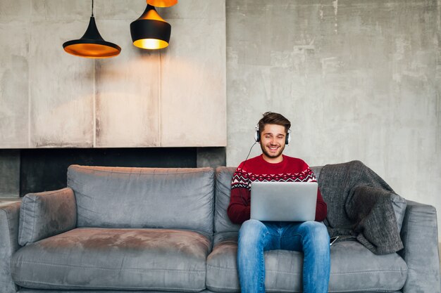 Young attractive man on sofa at home in winter in headphones, listening to music, wearing red knitted sweater, working on laptop, freelancer, smiling, happy, positive