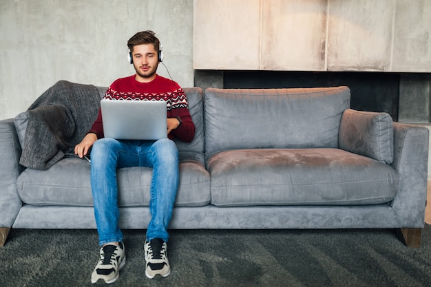 Giovane uomo attraente sul divano di casa in inverno in cuffia, ascoltando musica, indossando un maglione lavorato a maglia rosso, lavorando sul computer portatile, libero professionista