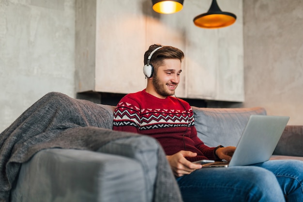 Young attractive man on sofa at home in winter with smartphone in headphones, listening to music, wearing red knitted sweater, working on laptop, freelancer, smiling, happy, positive, typing