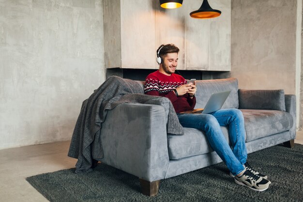 Young attractive man on sofa at home in winter with smartphone in headphones, listening to music, wearing red knitted sweater, working on laptop, freelancer, smiling, happy, positive