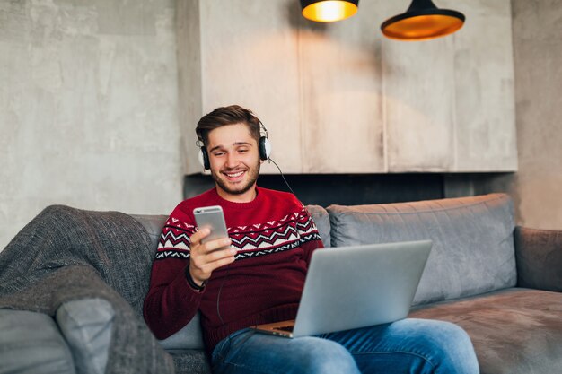 Young attractive man on sofa at home in winter with smartphone in headphones, listening to music, wearing red knitted sweater, working on laptop, freelancer, smiling, happy, positive