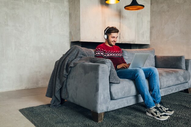 Young attractive man on sofa at home in winter with smartphone in headphones, listening to music, wearing red knitted sweater, working on laptop, freelancer