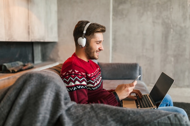 Giovane uomo attraente sul divano a casa in inverno con lo smartphone in cuffia, ascoltando musica, indossando un maglione lavorato a maglia rosso, lavorando sul computer portatile, libero professionista