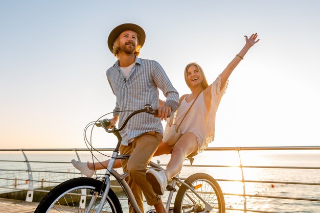 若い魅力的な男と女の自転車、日没で海で夏休みにロマンチックなカップル、自由奔放に生きる流行に敏感なスタイルの服、一緒に楽しんで友達