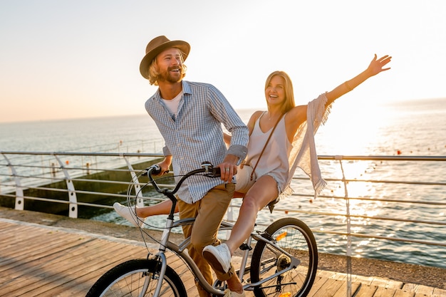 若い魅力的な男と女の自転車、日没で海で夏休みにロマンチックなカップル、自由奔放に生きる流行に敏感なスタイルの服、一緒に楽しんで友達