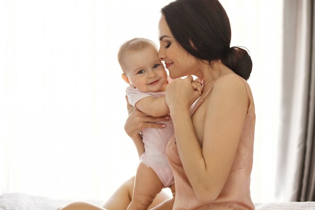 Foto gratuita giovane mamma attraente in indumenti da notte sorridente che abbraccia baciando il suo bambino seduto nel letto sopra la finestra occhi chiusi.