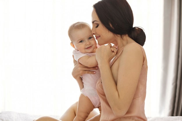 Young attractive mom in sleepwear smiling hugging kissing her baby sitting in bed over window Closed eyes.