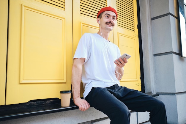 Young attractive mustache man in T-shirt and red hat listening music on cellphone while  happily looking aside with yellow background on city street