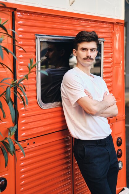 Молодой привлекательный усатый мужчина в белой футболке опирается на красный фургон, держась за руки, задумчиво глядя в камеру на городской улице